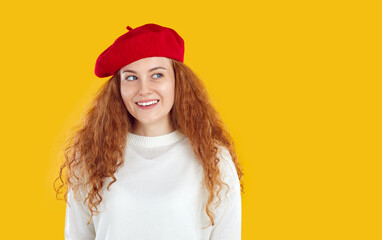 Portrait of beautiful happy girl with red wavy long hair, dressed in warm white jumper and red beret, looks away and smiles with snow-white toothy smile. Studio photo on isolated yellow background.