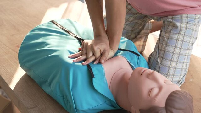 Close-up of young Asian female hands performing cardiopulmonary resuscitation (CPR) on a training dummy. Key elements include defibrillator, compressions, mouth-to-mouth, and abdominal thrusts.
