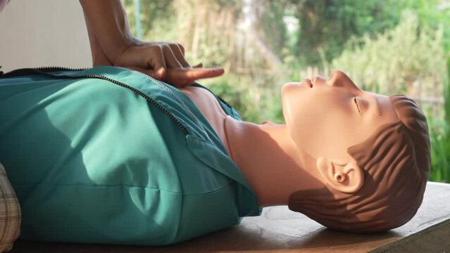 Close-up of young Asian female hands performing cardiopulmonary resuscitation (CPR) on a training dummy. Key elements include defibrillator, compressions, mouth-to-mouth, and abdominal thrusts.