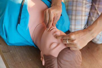 Close-up of young Asian female hands performing cardiopulmonary resuscitation (CPR) on a training dummy. Key elements include defibrillator, compressions, mouth-to-mouth, and abdominal thrusts.