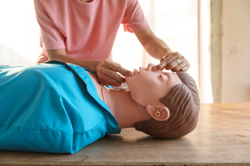 Close-up of young Asian female hands performing cardiopulmonary resuscitation (CPR) on a training dummy. Key elements include defibrillator, compressions, mouth-to-mouth, and abdominal thrusts.