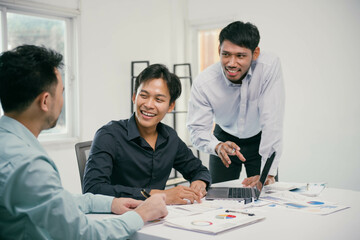 Three men are sitting at a table, one of them is pointing at a laptop