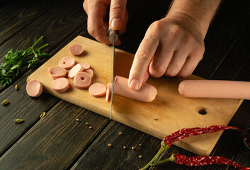 Slicing vienna sausage on a cutting board with a knife in a cook hand for preparing a dish for lunch. Space for menu on dark background - obrazy, fototapety, plakaty