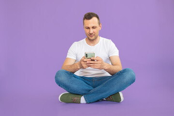 Young handsome Caucasian man sitting with mobile phone against violet background