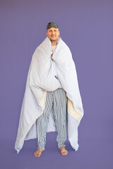 Full size body length young fun man 30s wear pajamas jam sleep mask resting relax at home wrap cover with blanket duvet look camera isolated on blue background Good mood night bedtime concept.