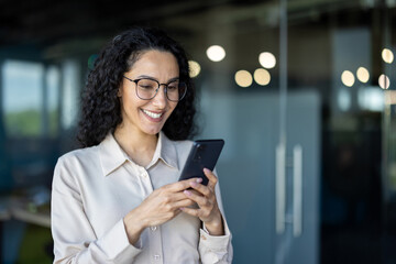 Beautiful woman in the office, happy and smiling latin american business woman uses internet phone close up, female worker reads message and browses internet pages inside office building .