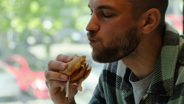 Junk street food concept. Close up young bearded man eating hamburger sitting in cafe. The guy enjoys biting into a burger with chicken cutlet, vegetables and bacon