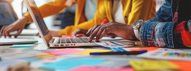 Close up of hands working on a laptop, with colorful stickers and markers on a desk with papers in an office environment