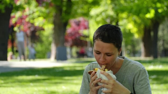 Young woman with short hair eats a gyros in summer park. Traditional Greek street food - chicken gyros with sauce, vegetables and French fries wrapped in pita. Middle eastern street food cuisine