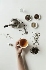 Elegant Hand Reaching For Tea Cup Amidst Assorted Leaves on White Table - Studio Captured, Natural Light