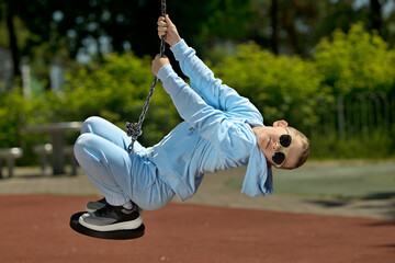 A little boy swinging on a swing.A boy in a blue tracksuit and sunglasses plays on the playground.