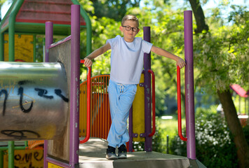 A little boy in a white T-shirt and glasses. A boy on a playground for children. A cheerful boy. Baby model. © Robert