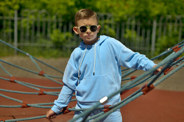 A little boy in a blue tracksuit and sunglasses. A boy on the playground. A cheerful boy. Baby model.