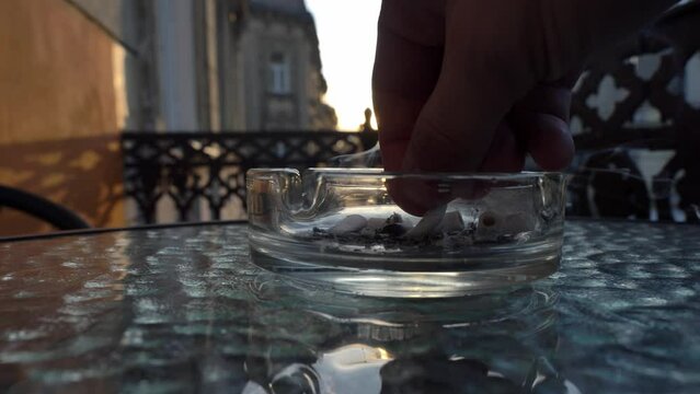 A man smokes and puts out the stub of a cigarette on an ashtray, sitting on the balcony in the evening during sunset. Close-up of an ashtray into which a man dumps cigarette ash while smoking.