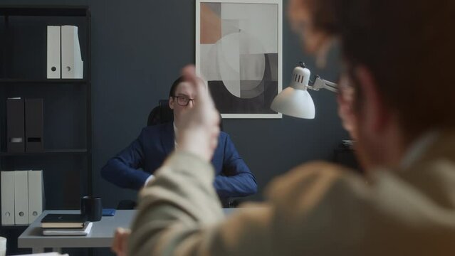 Medium shot of young Caucasian businessman arriving for work in morning, sitting at desk, taking out things and giving cold looks to buffoon colleague, who is having fun with wooden toys to annoy him