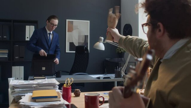 Medium pan shot of red-haired Caucasian guy sitting in office in morning, playing guitar to harrass male colleague in business suit arriving for work, clowning about and waving at him with wooden hand