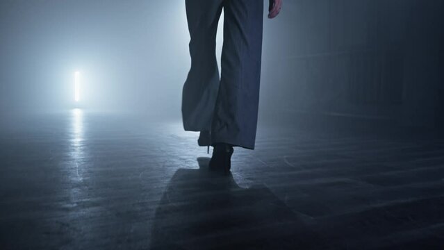 Dancer legs walking through dark studio. Young woman entering the stage with smoke and lights on background. Young woman walking in high heels. Dance concept. Tracking shot in 4K, UHD