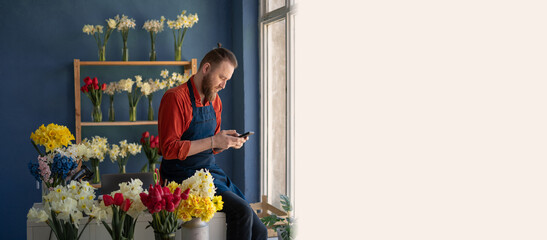 male self employed florist sitting at work desk focused on mobile phone screen. Man flower arranger...