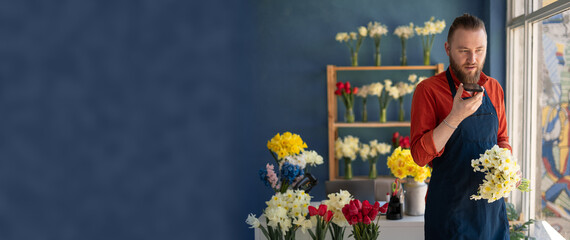 Male florist recording audio message on smartphone, chatting with client online, giving consultation standing in his floral shop