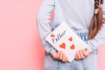 Little girl holding behind her back a drawn card with hearts for Father's Day holiday in front of a pink background.