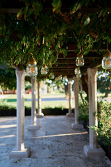 Lamps in glass jars hang from the beams of a long pillared pergola in a garden