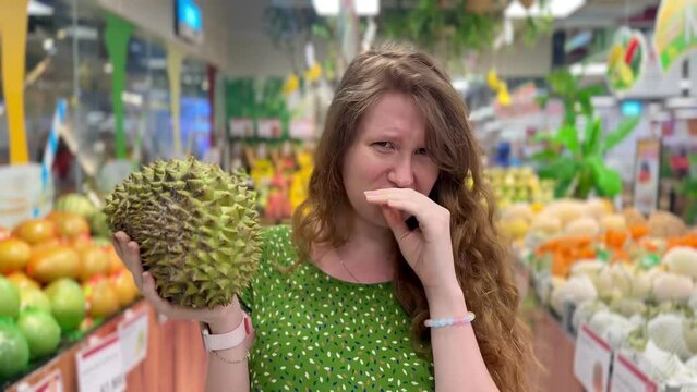 a woman in a store holds the tropical fruit durian and winces from the pungent smell
