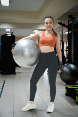 A woman is holding an exercise ball in a gym. She is smiling and she is enjoying her workout.