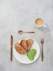 Top view of white plate with slices of toasted bread and sliced avocado in fan shape. Bronze colored coffee cup and silverware on gray table with copy space.