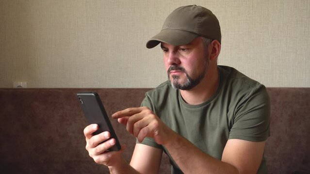 A man with a beard. Holding a black smartphone in his hand. Presses his finger on the screen. Thoughtful look. Green baseball cap and T-shirt. Military. In room. Sitting on a brown sofa against the ba