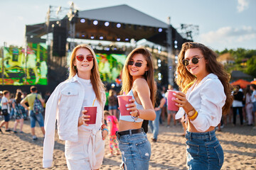 Group of girls at summer beach music festival, raising glasses in toast, enjoying sunny day with live concert in background. Smiling females celebrate, dance with drinks in hand.
