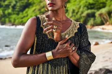 Flamboyant queer black male with brass jewelry poses on scenic ocean beach. Trans sexual ethnic...