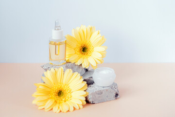 Cosmetic serum bottle and cream jar on stone podium with yellow gerbera flowers on beige table....