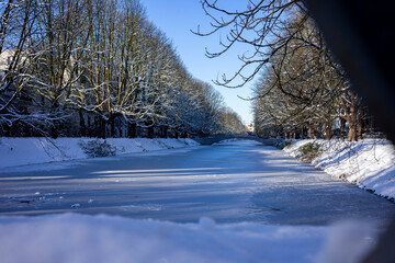 Winter's Grip on Clarenbachkanal, Cologne: Snow-dusted trees line the frozen canal under a clear...