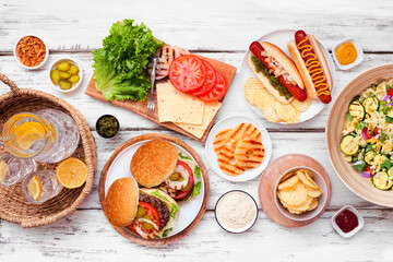 Summer BBQ or picnic table scene with hamburgers, hotdogs, salad and snacks. Top down view over a white wood background.