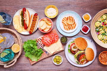 Summer BBQ or picnic table scene with hamburgers, hotdogs, salad and snacks. Top view over a dark wood background.