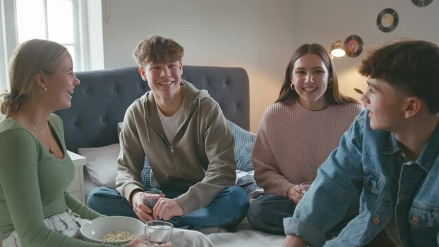 Group of teenage girls and boys at home hanging out laughing and talking in bedroom together with soft drinks and popcorn - shot in slow motion