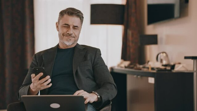 Content middle-aged man multitasking with phone and laptop. Businessman working at home with computer, smiling. Happy mid adult male with content smile. Entrepreneur managing business online.