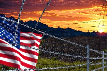 A red, white, and blue American flag is flying in front of a barbed wire fence