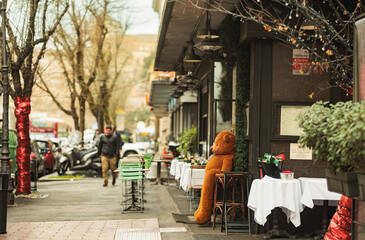 A cozy teddy bear sits on a wooden table outside a charming restaurant, surrounded by the bustle of...