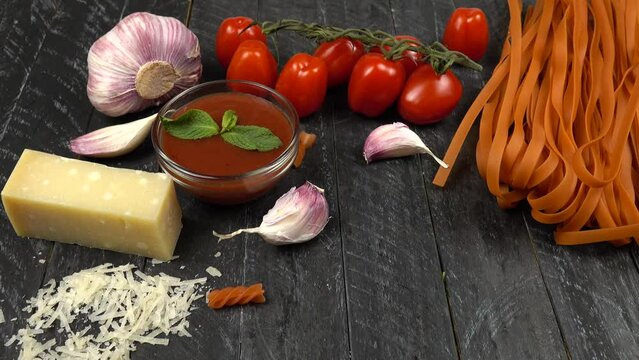 Italian cuisine, red pasta and ingredients for it.  Cherry tomatoes, garlic, parmesan cheese, red noodles and tomato sauce. Red  pasta  fusill falling on a black cutting board.