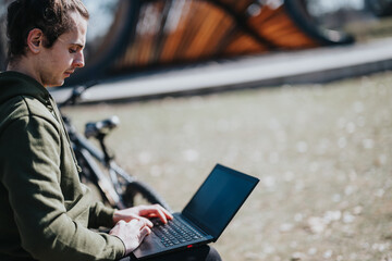 A focused young male works on his laptop seated beside his bicycle outdoors, enjoying the sunny...