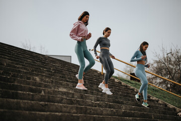 Three athletic women in workout gear descend a long staircase while exercising in an urban outdoor...