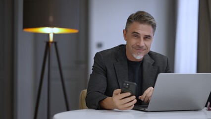 Professional man with cell phone and laptop in an office. Businessman working at home with computer, smiling. Happy mid adult male with content smile. Entrepreneur managing business online.