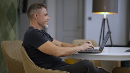 Relaxed mature man focused on work, typing on laptop indoor. Businessman working at home with computer, smiling. Happy mid adult male with content smile. Entrepreneur managing business online.