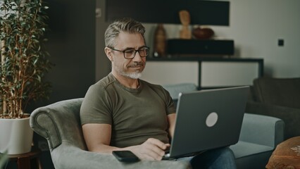 Man in 50s sitting in living room, working with laptop computer. Happy mid adult, mature aged casual male indoors, smiling. Businessman in home office managing business online.