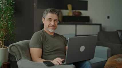 Confident mature man using laptop and smartphone, sitting in modern living room. Businessman working at home with computer, smiling. Happy mid adult man. Entrepreneur managing business online.