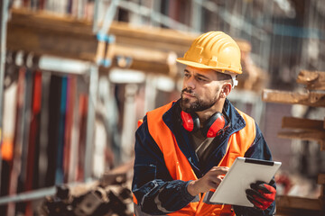 Construction site manager standing  wearing safety vest and helmet,using his tablet at construction...