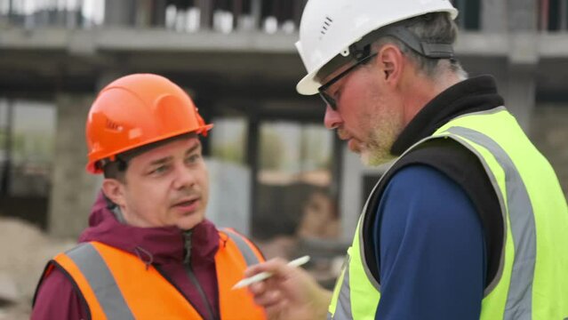 Worker in an orange helmet gives evidence of an accident on a construction site to an occupational safety inspector looking into a digital tablet.