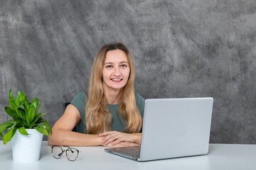 Young woman with blonde hair in gray dress posing in front of a laptop beside a green flower