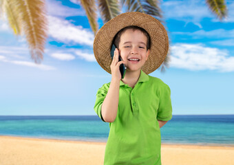 Little boy using smart phone with happy face and hat standing with confident smile showing teeth at tropical beach background